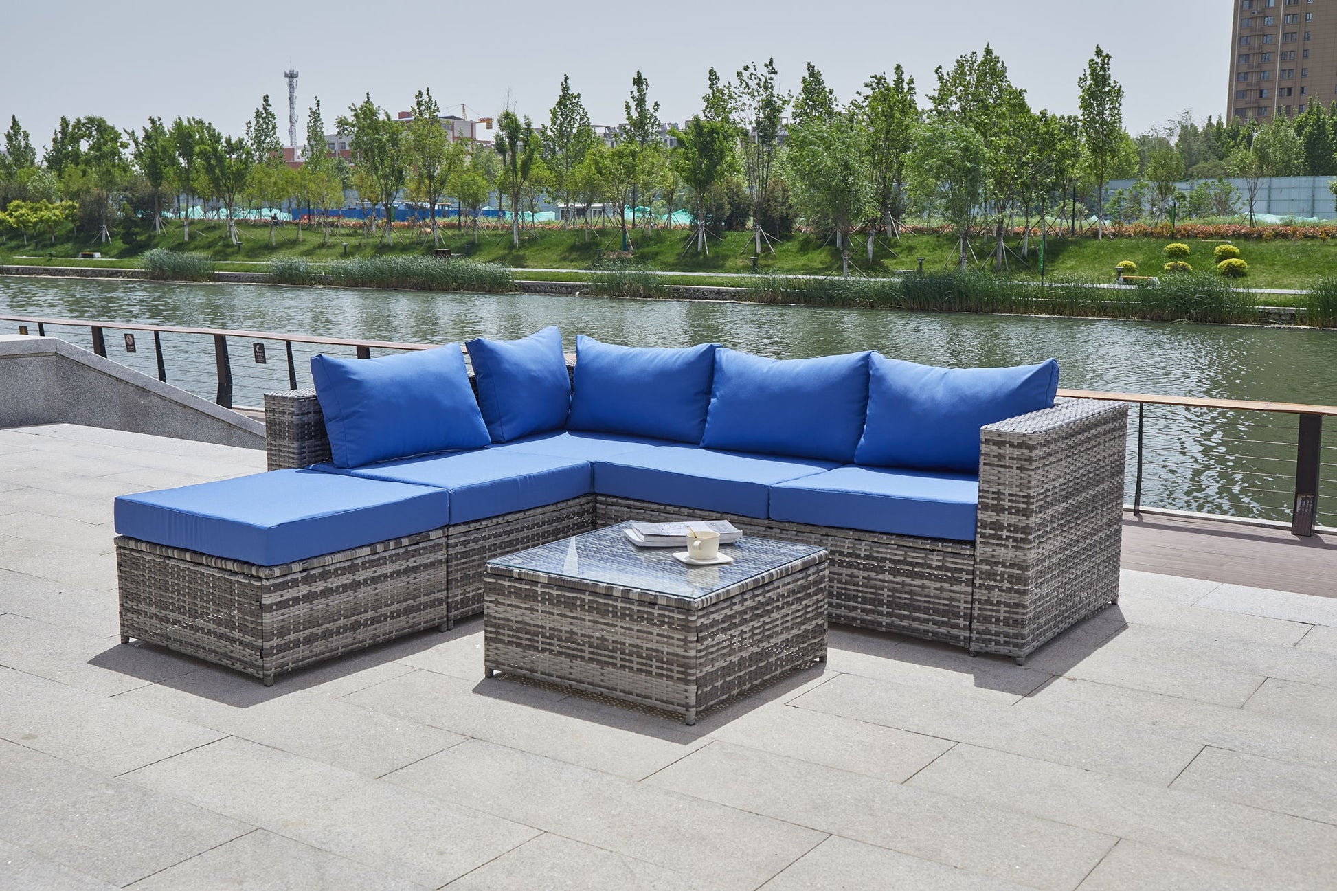 A Patio Sofa With Cushions featuring gray wicker and blue cushions is set around a glass-top coffee table on a stone terrace that overlooks a tree-lined river.