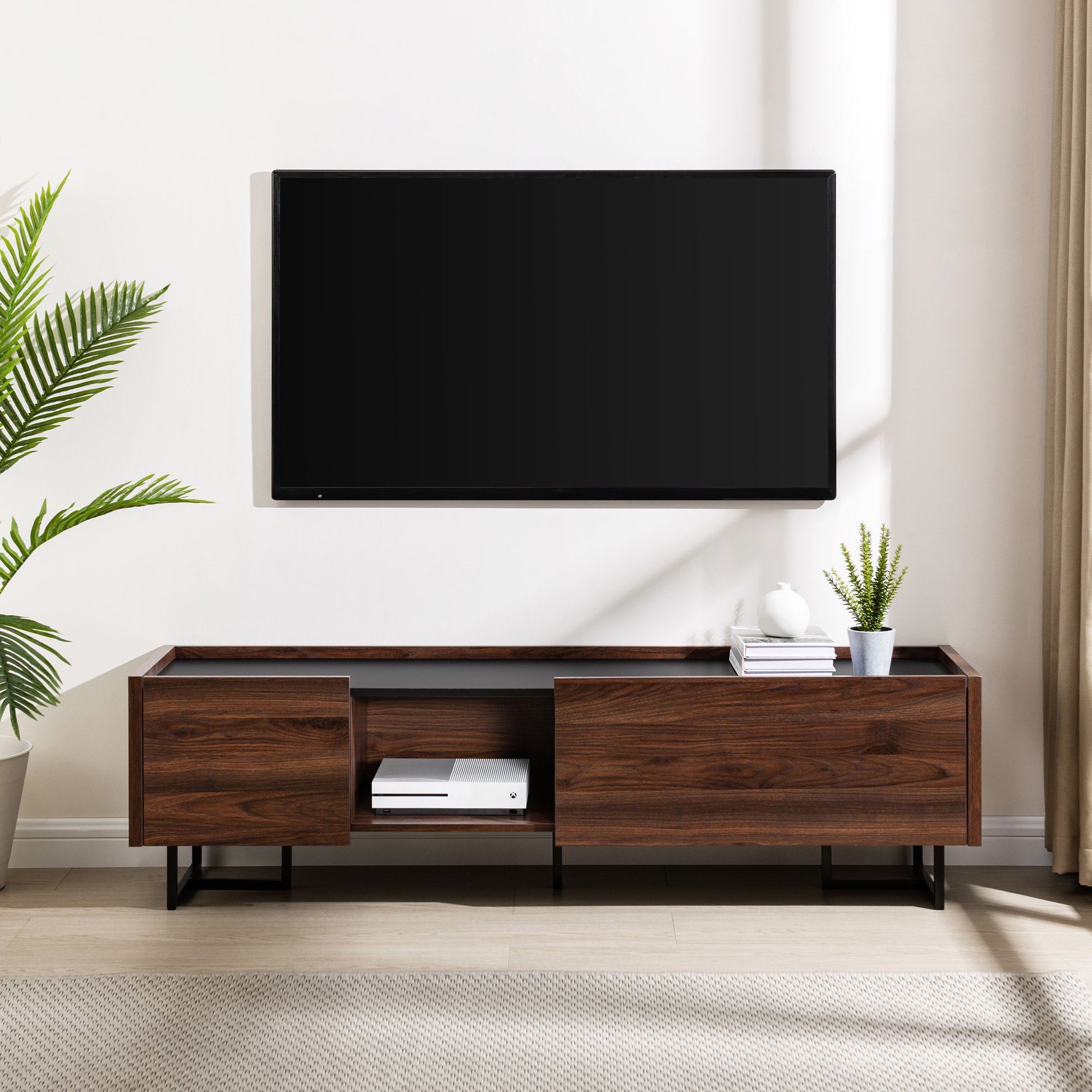 A wall-mounted flat-screen TV sits above a Contemporary Low Profile TV Stand, which holds plants, books, and a device. A large green plant is on the left, and beige curtains are positioned to the right.