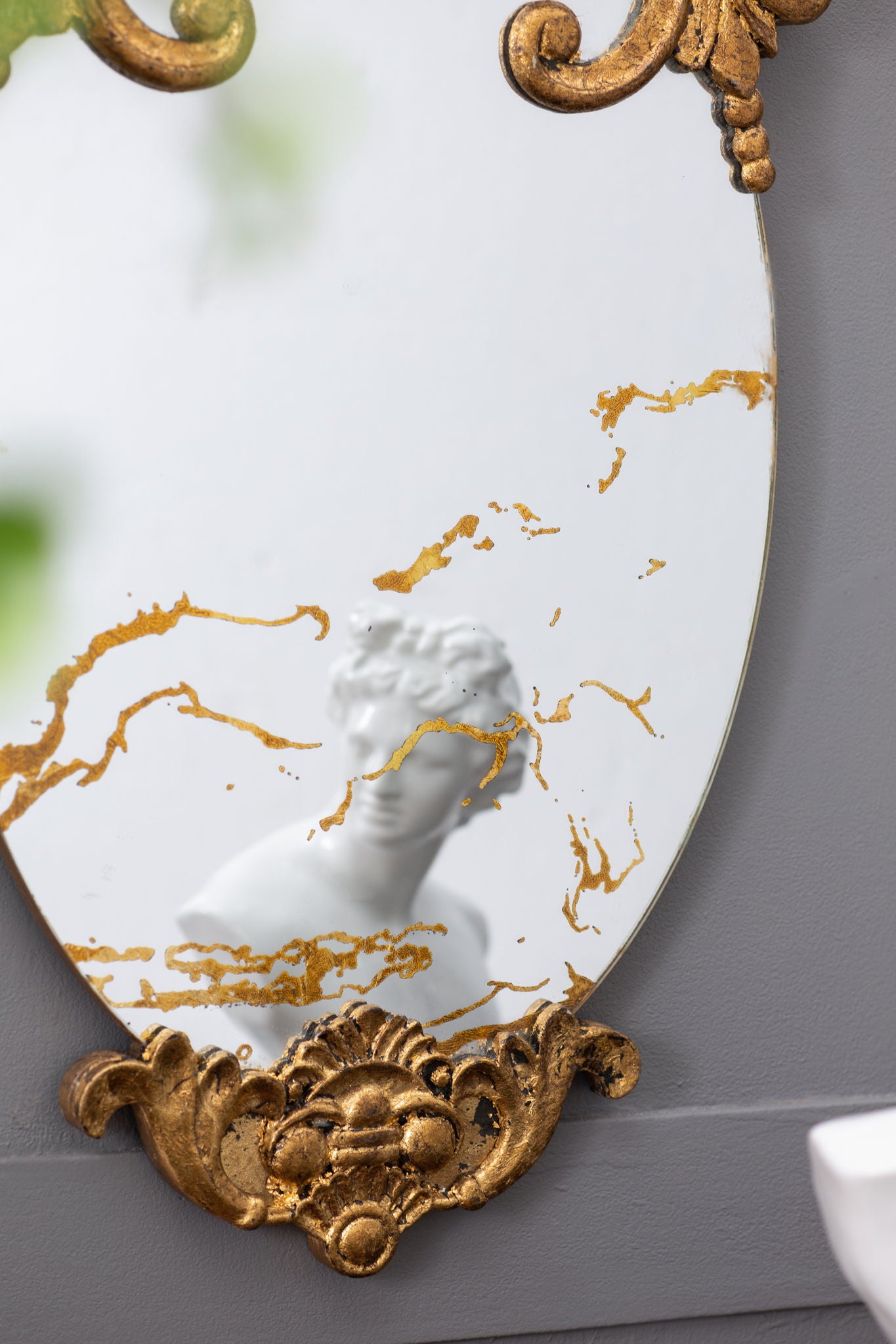 The Decorative Oval Wall Mirror, Accent Mirror for Living Room, Entryway, Bedroom features ornate gold patterns. Mounted on a gray wall, it beautifully reflects a white classical-style bust.