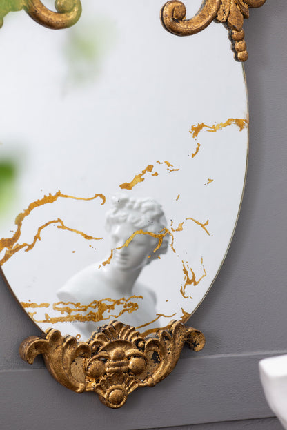 The Decorative Oval Wall Mirror, Accent Mirror for Living Room, Entryway, Bedroom features ornate gold patterns. Mounted on a gray wall, it beautifully reflects a white classical-style bust.