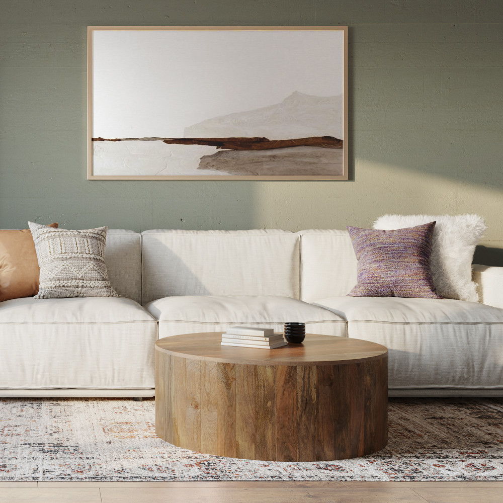 A modern living room with a white sofa, patterned cushions, the Millbury Drum Coffee Table in natural wood topped with books and a vase, a light patterned rug, and a minimalist landscape painting on a green wall.