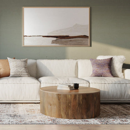 A modern living room with a white sofa, patterned cushions, the Millbury Drum Coffee Table in natural wood topped with books and a vase, a light patterned rug, and a minimalist landscape painting on a green wall.