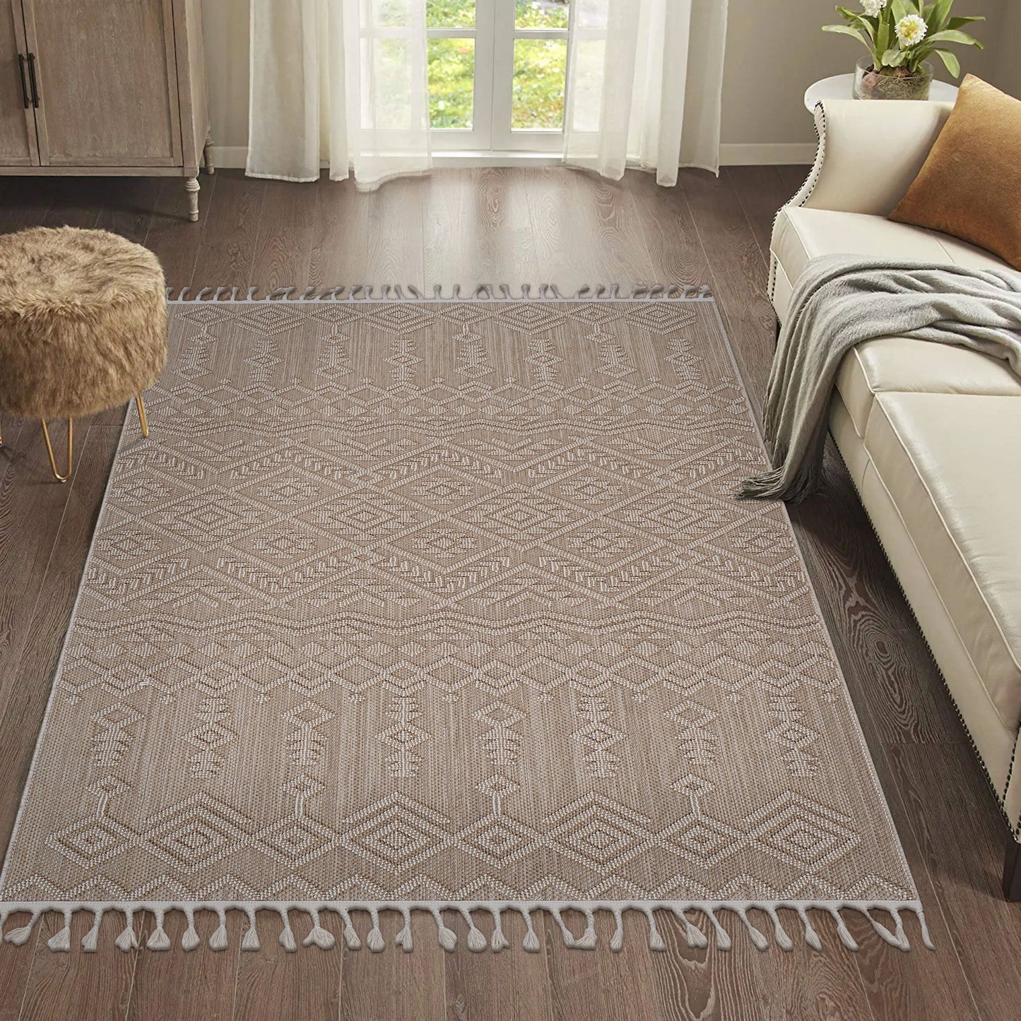 The Guros - Traditional Indoor / Outdoor Area Rug, featuring a beige pattern and tasseled edges, is laid on a dark wood floor in a bright living room with a cream sofa, brown furry ottoman, and sheer white curtains at the window.
