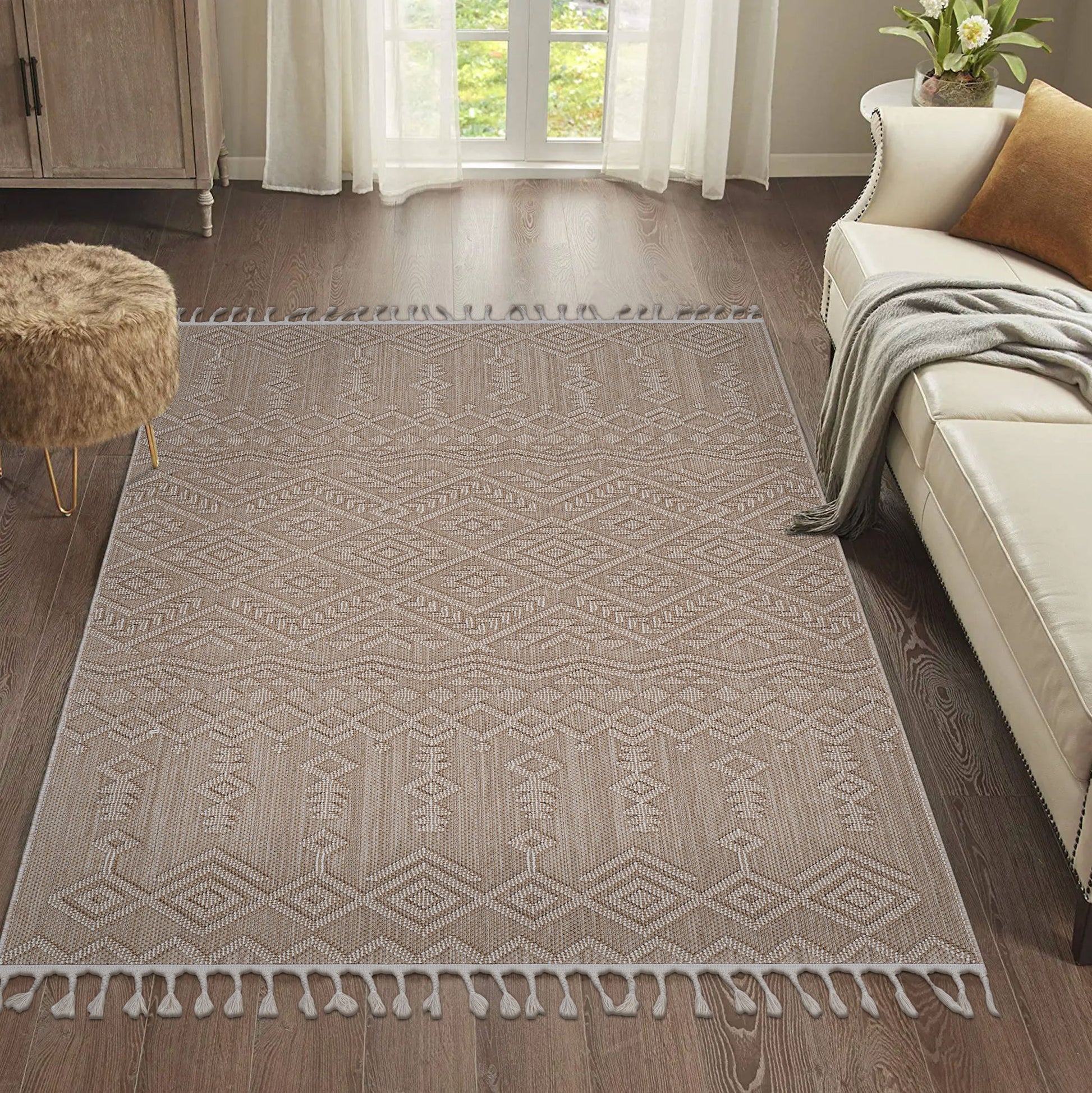The Guros - Traditional Indoor / Outdoor Area Rug, featuring a beige pattern and tasseled edges, is laid on a dark wood floor in a bright living room with a cream sofa, brown furry ottoman, and sheer white curtains at the window.