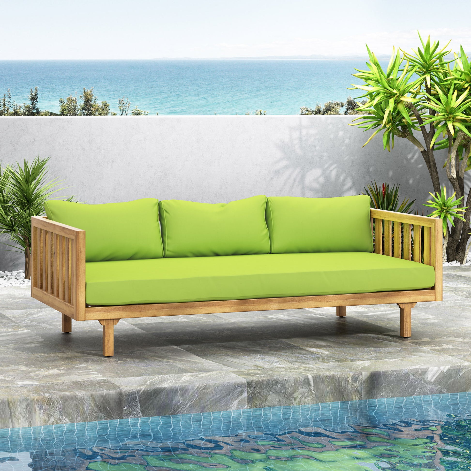 The Claremont 3 Seater Daybed, made of teak and acacia wood with water resistant green cushions, is set on a stone patio beside a pool, surrounded by tropical plants and an ocean view under a clear sky.