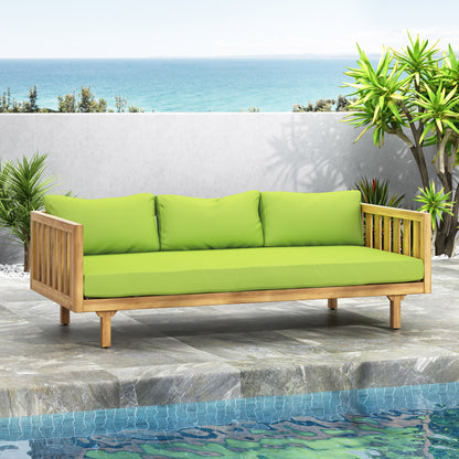 The Claremont 3 Seater Daybed, made of teak and acacia wood with water resistant green cushions, is set on a stone patio beside a pool, surrounded by tropical plants and an ocean view under a clear sky.
