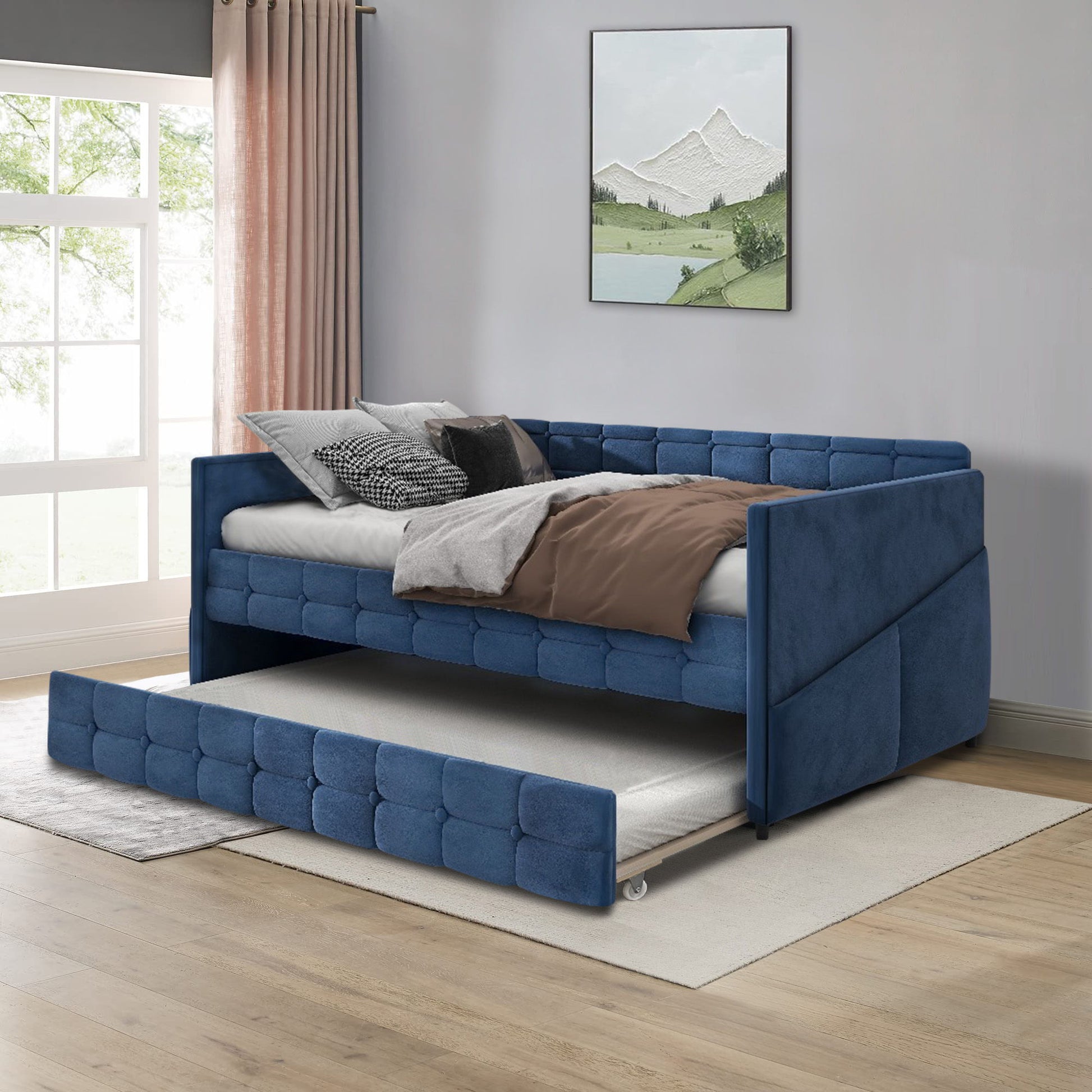 An Upholstered Twin Size Daybed Bed Frame with trundle and velvet fabric sits on a light rug in a bright room with wood floors, pink curtains, a large window, and a mountain landscape painting.