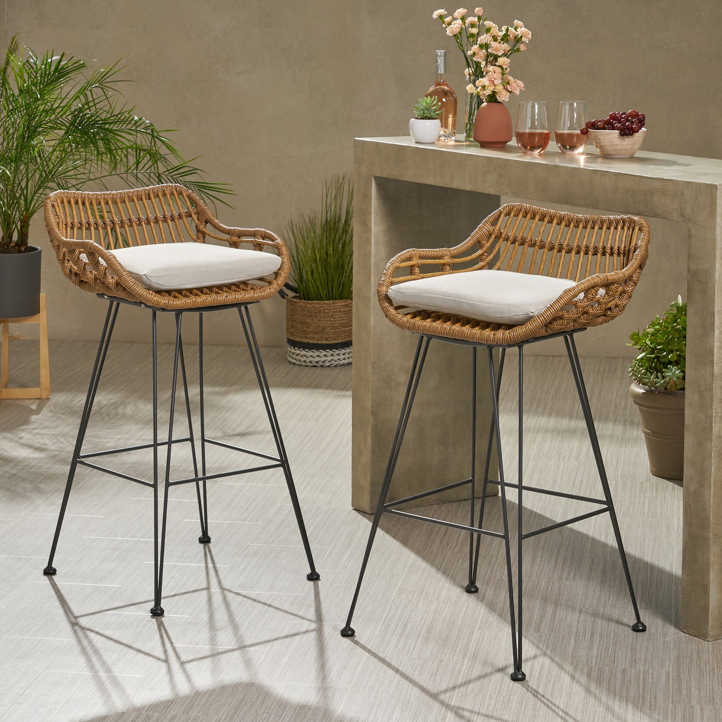 Two Outdoor Rattan Bar Chairs with metal legs and light cushions sit by a stone counter with drinks and flowers, while green potted plants accent the bright, modern indoor space.