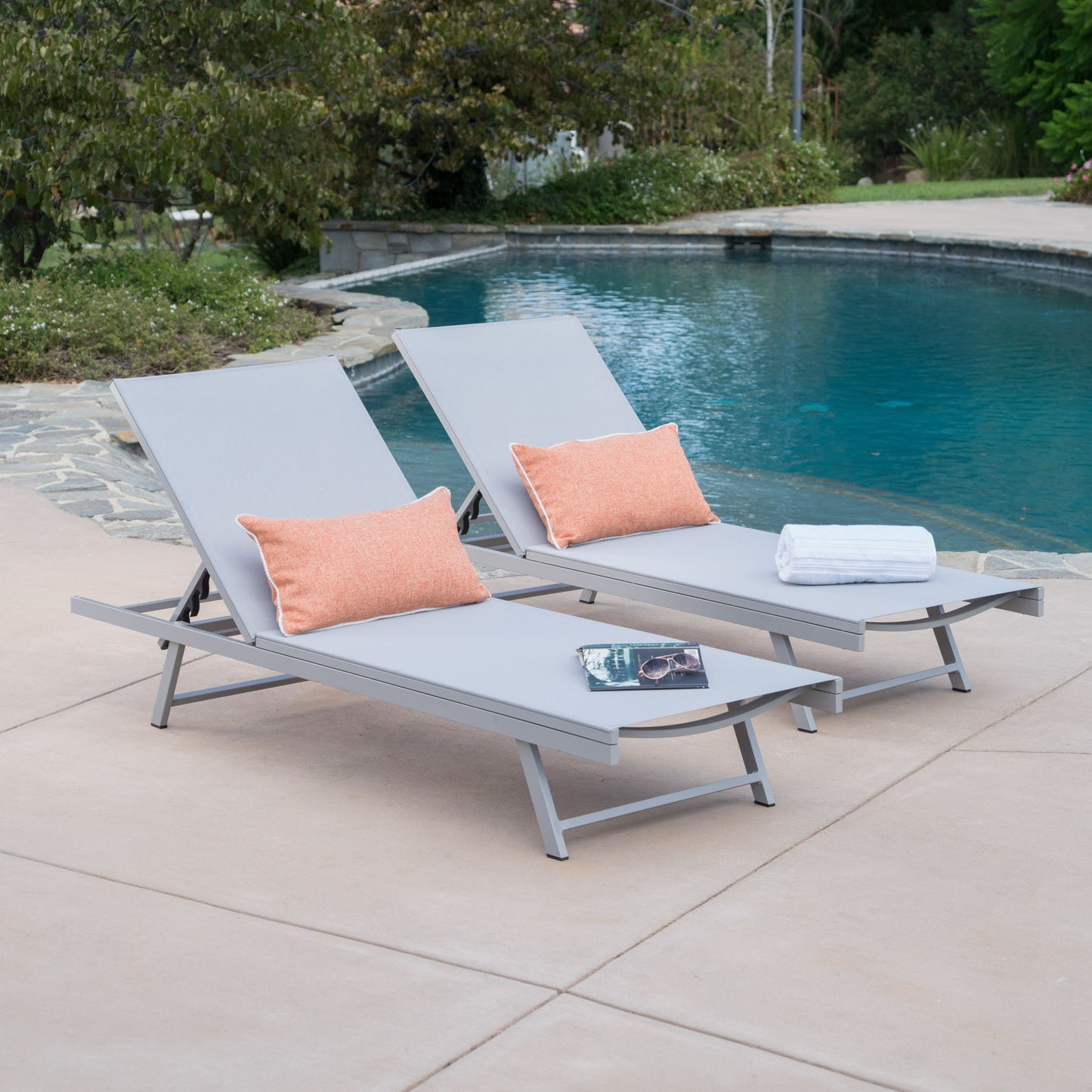Two Salton - Chaise Lounge Outdoor Relaxation chairs with orange pillows sit by a pool—one holds white towels, the other a book and sunglasses—surrounded by trees and landscaping.