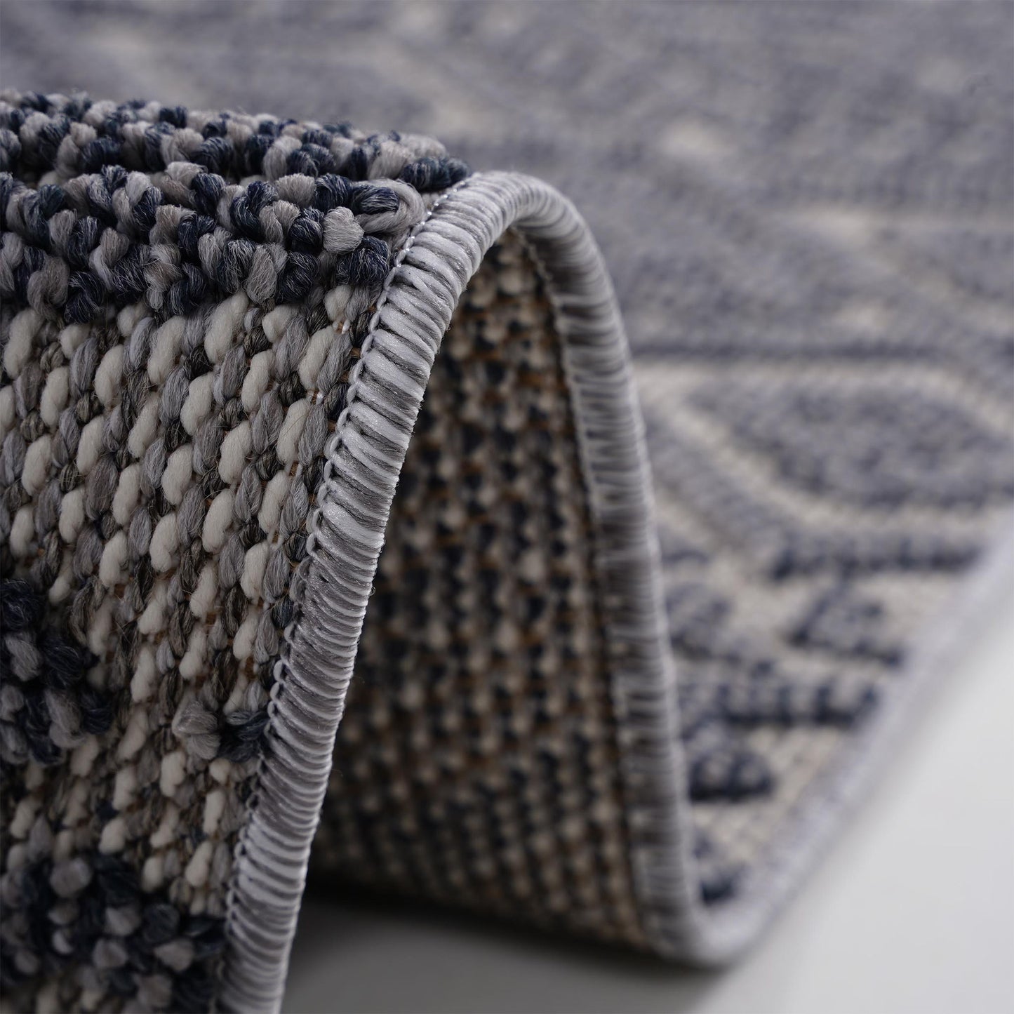 A close-up of the Guros - Traditional Indoor / Outdoor Area Rug, featuring a folded, textured surface with a woven gray and blue pattern and detailed edge stitching.