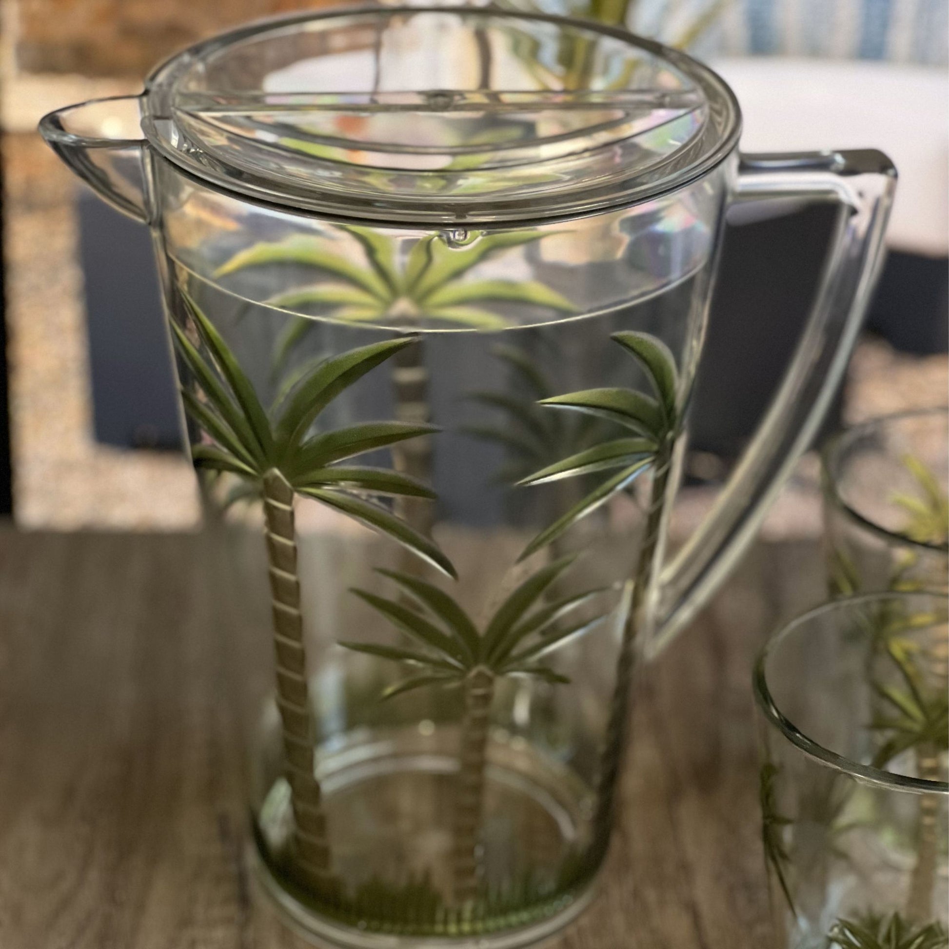 The Unbreakable Plastic Water Pitcher With Lid, Palm Tree Design - Clear sits on a wooden surface, featuring painted palm trees. Matching palm tree drinking glasses are placed nearby, all with sturdy handles and lids.