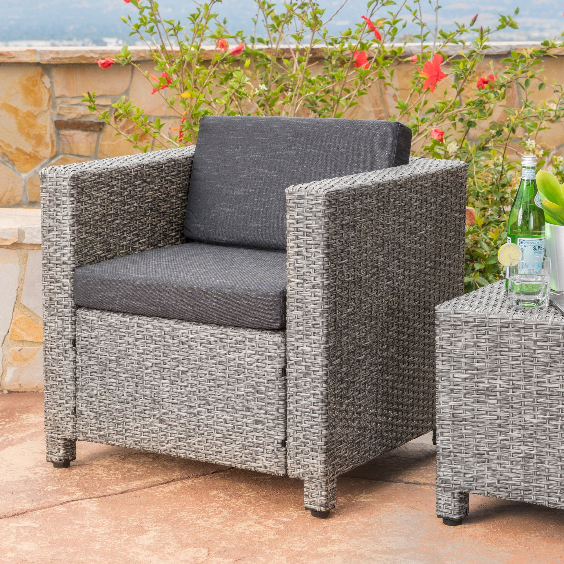 The Stylish Outdoor Club Chair with weather-resistant cushions in gray sits on a stone patio next to a matching table, with glass bottles and flowers displayed in the background.