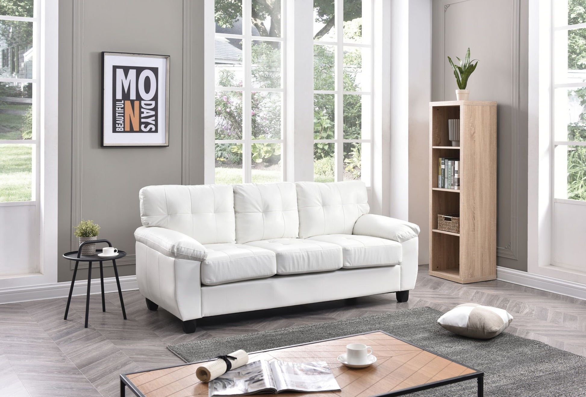 A bright, modern living room with large windows features the Comfortable Sofa in white, a wooden bookshelf with a plant, a side table holding a black cup, and a coffee table displaying an open magazine with a cushion on the floor.
