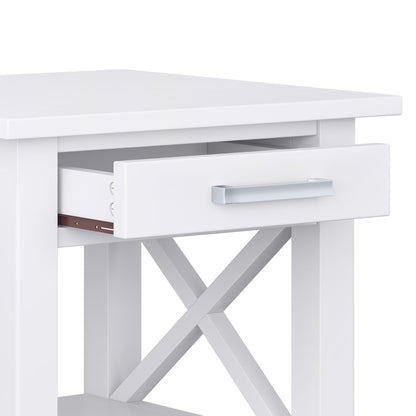 A close-up of the Kitchener - Handcrafted Table, featuring a white finish, open drawer with a silver handle, X-shaped side crossbars for support, a flat tabletop, and an open lower shelf for extra storage.