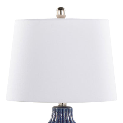 A close-up of the Rockwell - Contemporary Table Lamp (Set of 2), featuring a white cylindrical lampshade with a silver finial and a textured blue base partially visible.