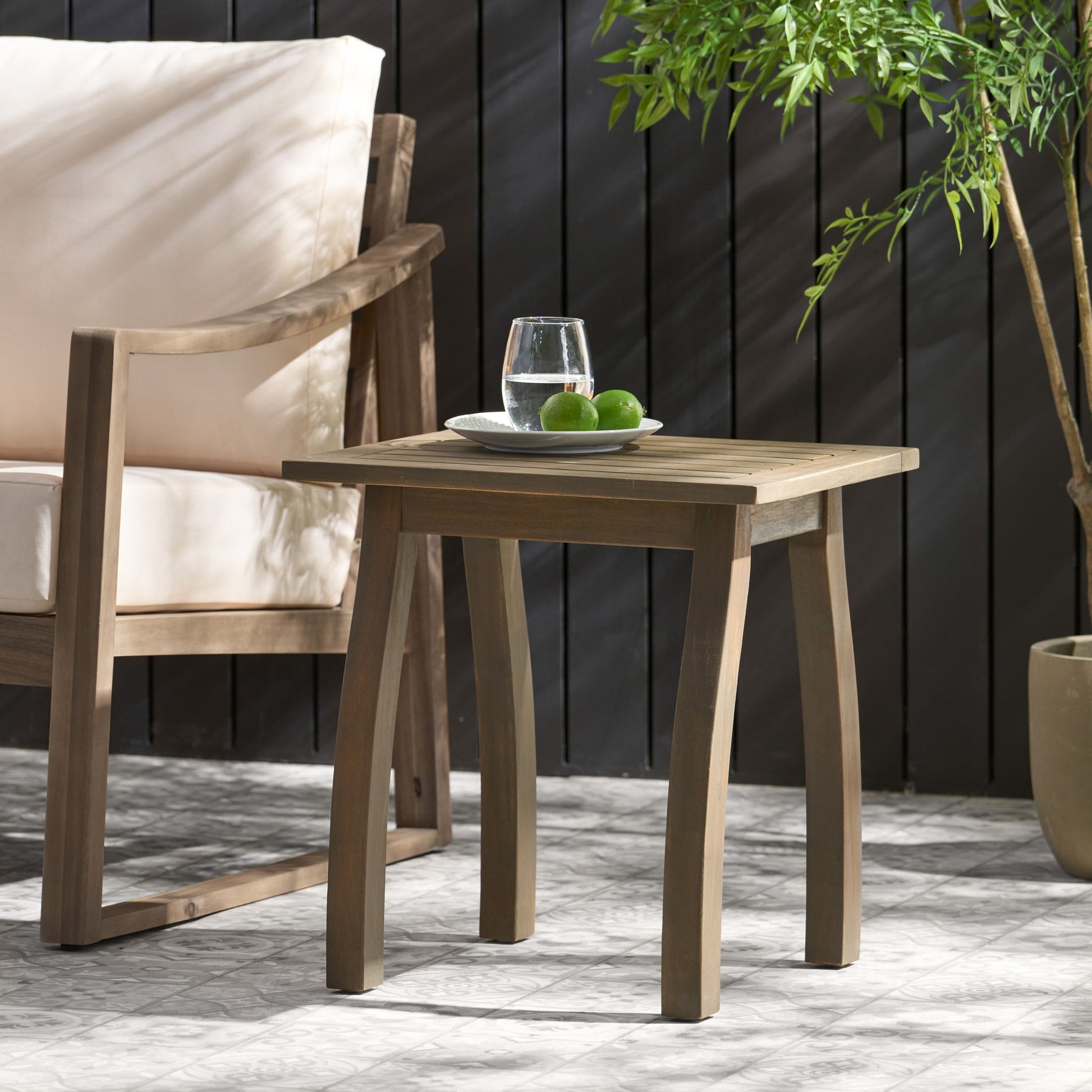 The Kinesis Outdoor Side Table with a slatted top and wood frame holds a plate of limes and a glass of water, set beside a cushioned wooden chair and a potted plant near a dark fence.