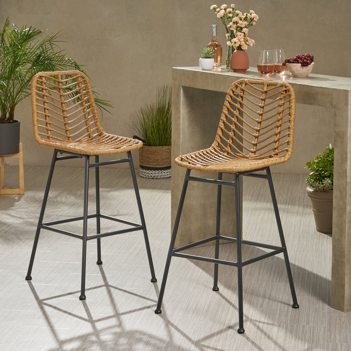 Two Sawtelle Outdoor Wicker Barstools (Set of 2) with black metal legs are placed by a beige counter holding flowers, wine glasses filled with pink drinks, a wine bottle, and a bowl of grapes. Green potted plants sit nearby.