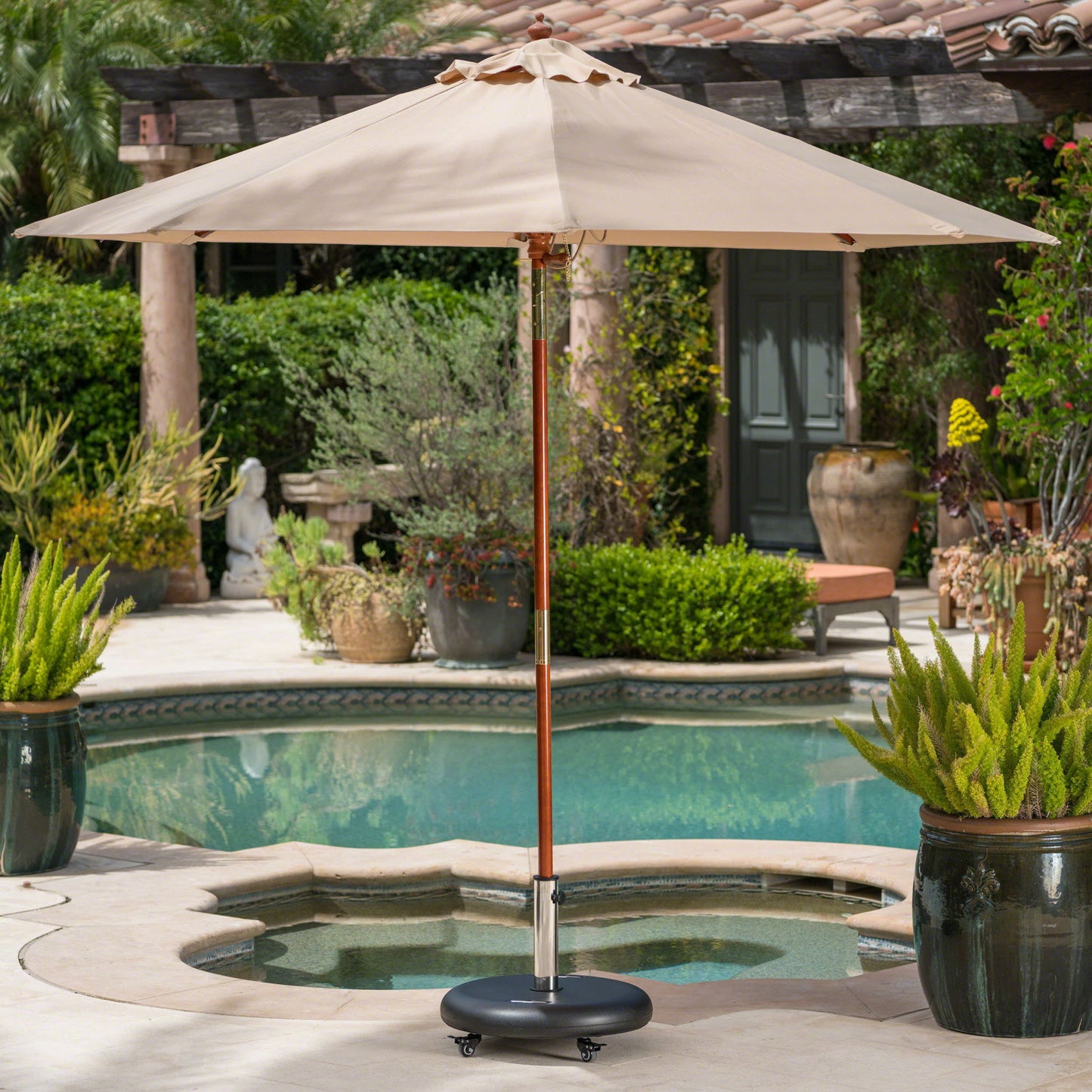 A beige patio umbrella with a wooden pole sits in a courtyard by a small round pool, supported by the Wheelie Round Umbrella Base With Stainless Steel Holder, surrounded by potted plants and outdoor greenery.