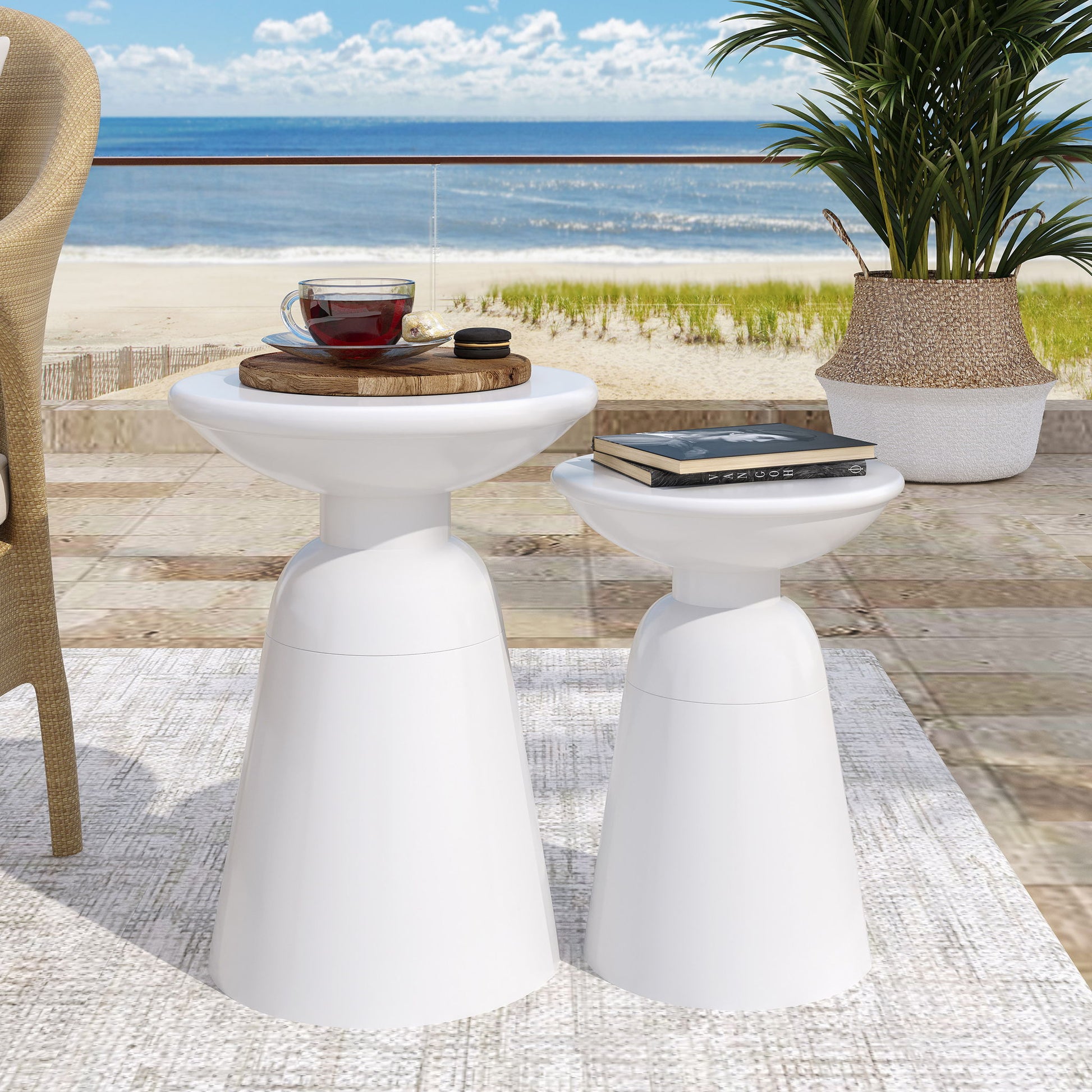 Two Serenita Outdoor Side Tables with flared design sit on a beach patio—one holds a wooden tray with glass teapot and cup, the other a closed book. A potted plant, plus sand, sea, and sky fill the serene background.
