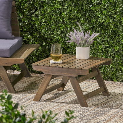 The Sedona Outdoor Side Table with open slat design holds a drink and potted lavender on a patterned rug next to a cushioned chair, set before a green hedge.
