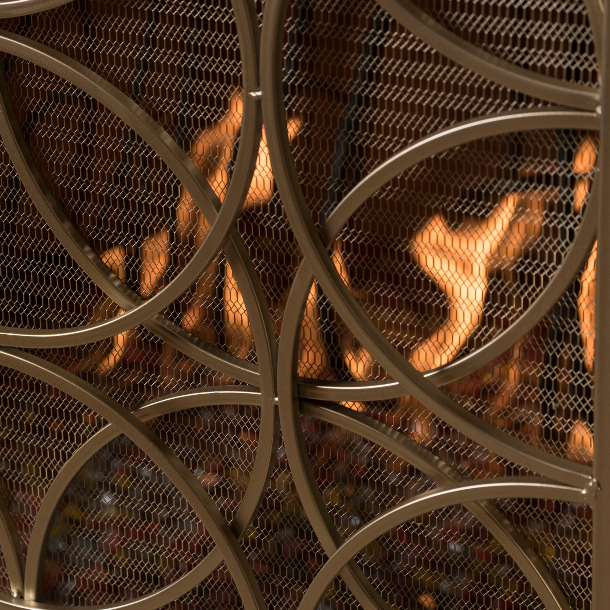 Close-up of the Thayer Single Panel Iron Fireplace Screen featuring a geometric mesh design, positioned in front of a fireplace with visible orange flames in the background.
