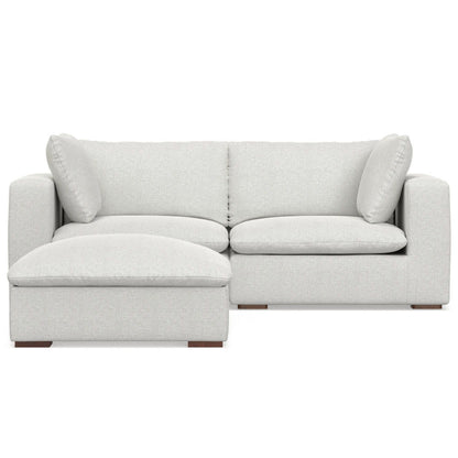 The Jasmine - Handcrafted Sofa And Ottoman is a light gray L-shaped sectional with wide armrests, two back cushions, two side pillows, an extended left chaise for lounging, and short block-style wooden legs.