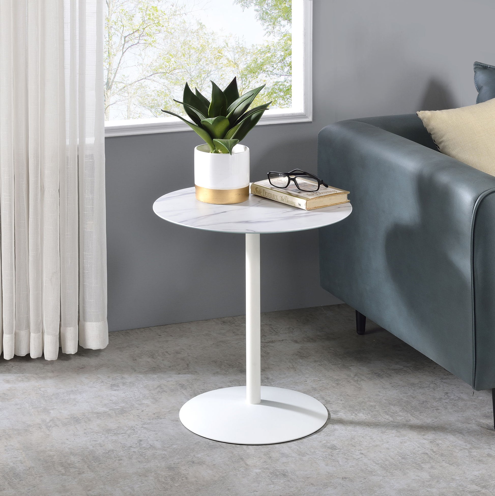 The Circa 17.5" End Table With Marble Textured Top holds a potted plant, eyeglasses, and a closed book beside a dark green sofa near a window with sheer curtains.