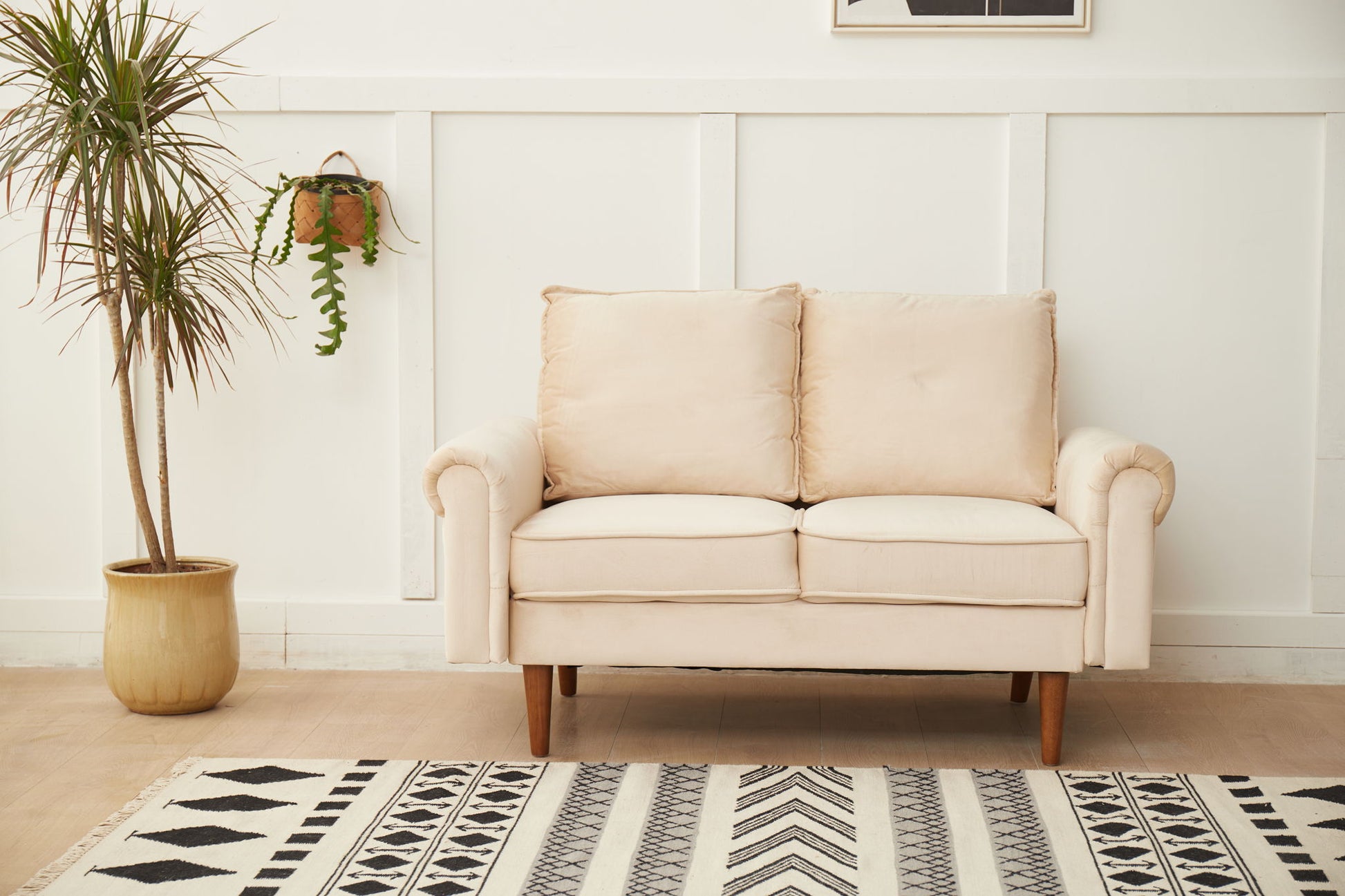 The Graceful Design Plush Comfort Loveseat in beige is set against a white paneled wall, with a potted plant to the left and a hanging fern nearby. A black and white patterned rug lies on the floor in front of the loveseat.