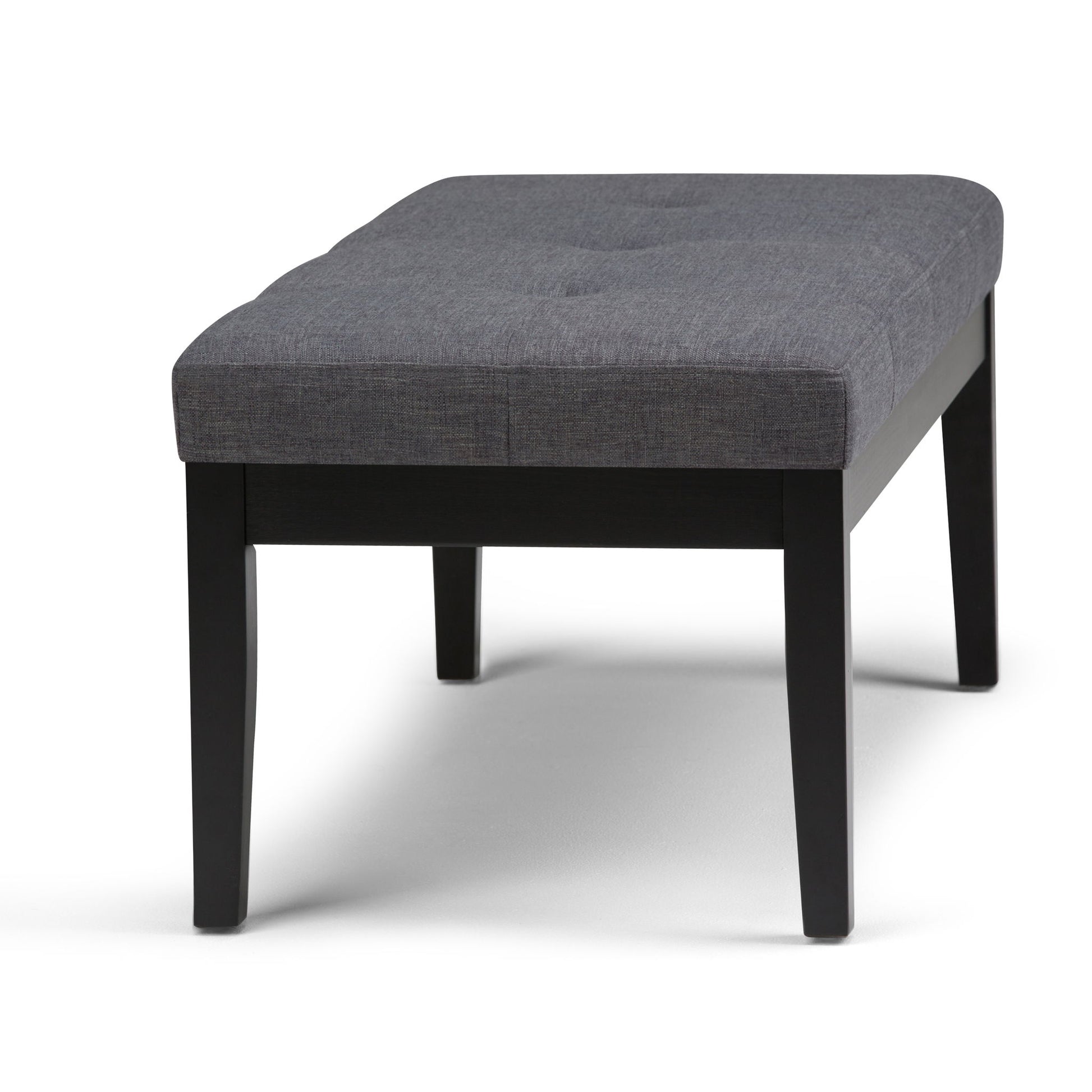 The Lacey Upholstered Tufted Ottoman Bench features a cushioned gray fabric seat with subtle tufted detailing and sturdy black wooden legs.