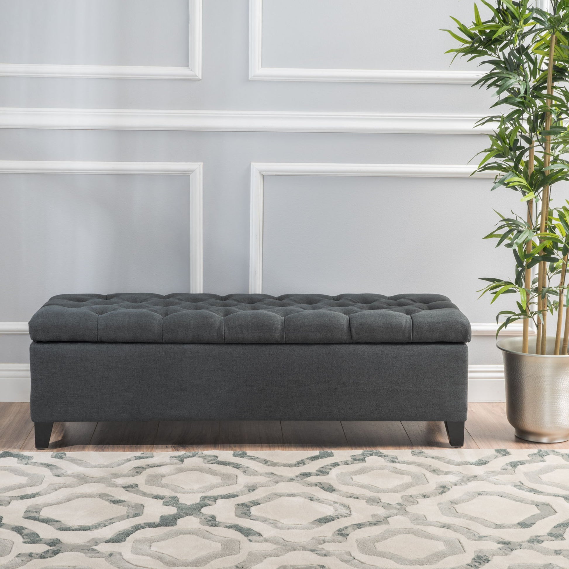 The Glouster Contemporary Storage Ottoman with a button-tufted design sits on a patterned rug beside a potted plant, set against a light gray wall with white trim and molding.