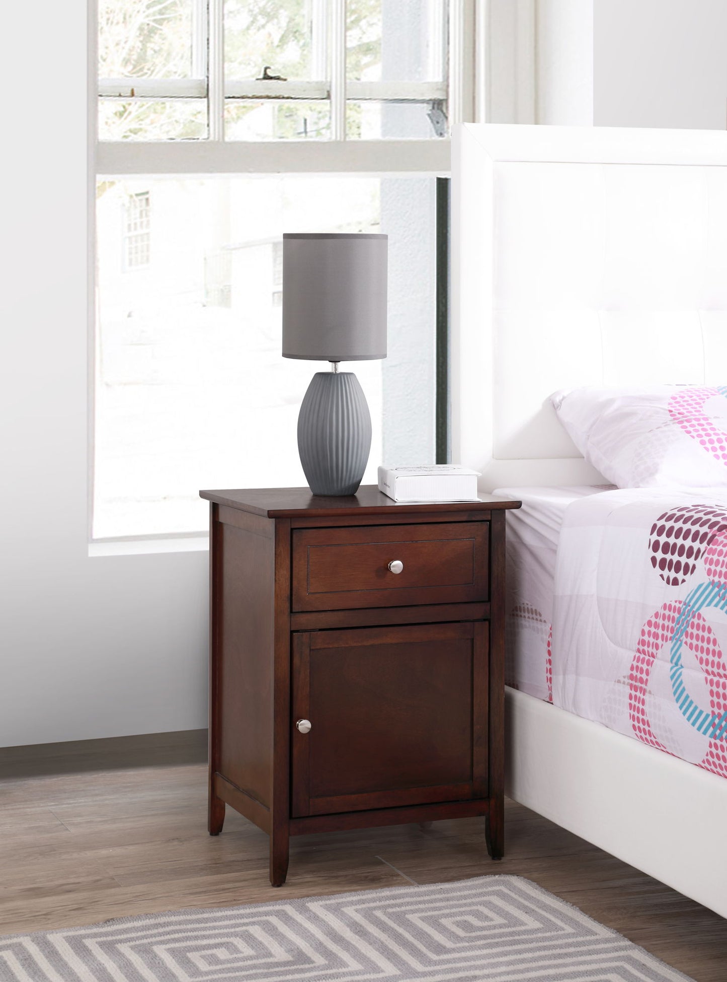 The Izzy - 1 Drawer /1 Door Nightstand stands by a white bed. It features a drawer and cabinet, topped with a gray lamp and tissue box. A window behind it bathes the scene in natural light.