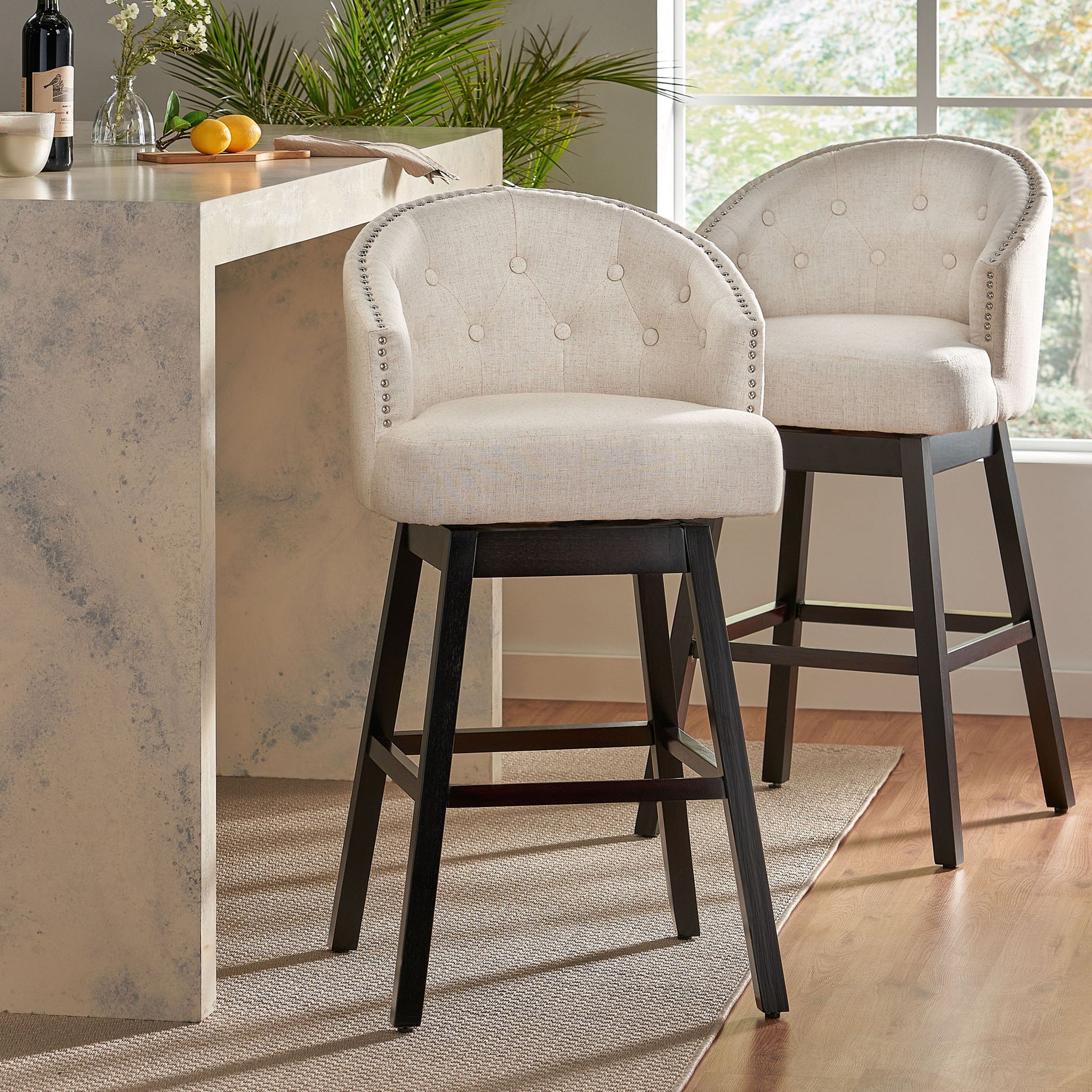 Two Mellifluous 360 Degree Swivel Bar Stools with light beige upholstery, button tufting, nailhead trim, and dark wooden legs sit at a modern marble kitchen island, bathed in natural light from a nearby window.