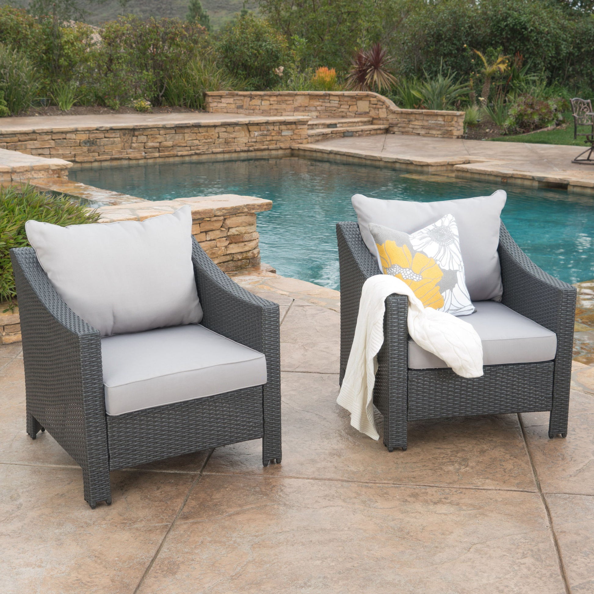 Two Antibes armchairs with water resistant cushions are on a stone patio by the pool; one features a floral pillow and a white blanket. Lush greenery and landscaped stone walls provide a scenic backdrop.