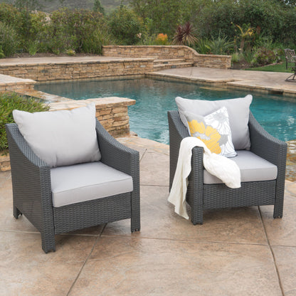 Two Antibes armchairs with water resistant cushions are on a stone patio by the pool; one features a floral pillow and a white blanket. Lush greenery and landscaped stone walls provide a scenic backdrop.