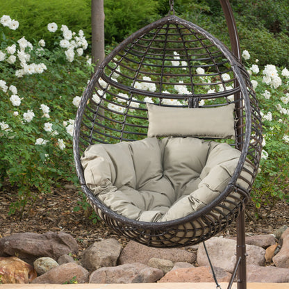 The Hanging Basket Chair With Metal Frame And Seat Cushion, featuring a beige pillow, is placed outdoors in a garden among rocks, mulch, lush green plants, and blooming white flowers.