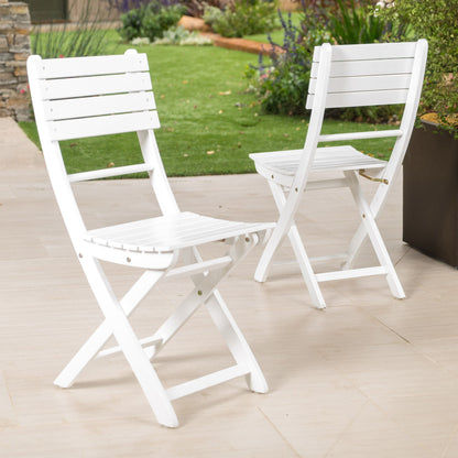 Two Outdoor Foldable Wood Dining Chairs With Slat Design (Set of 2) are arranged on a light patio, with green grass, plants, and a flower bed in the background for a garden ambiance.
