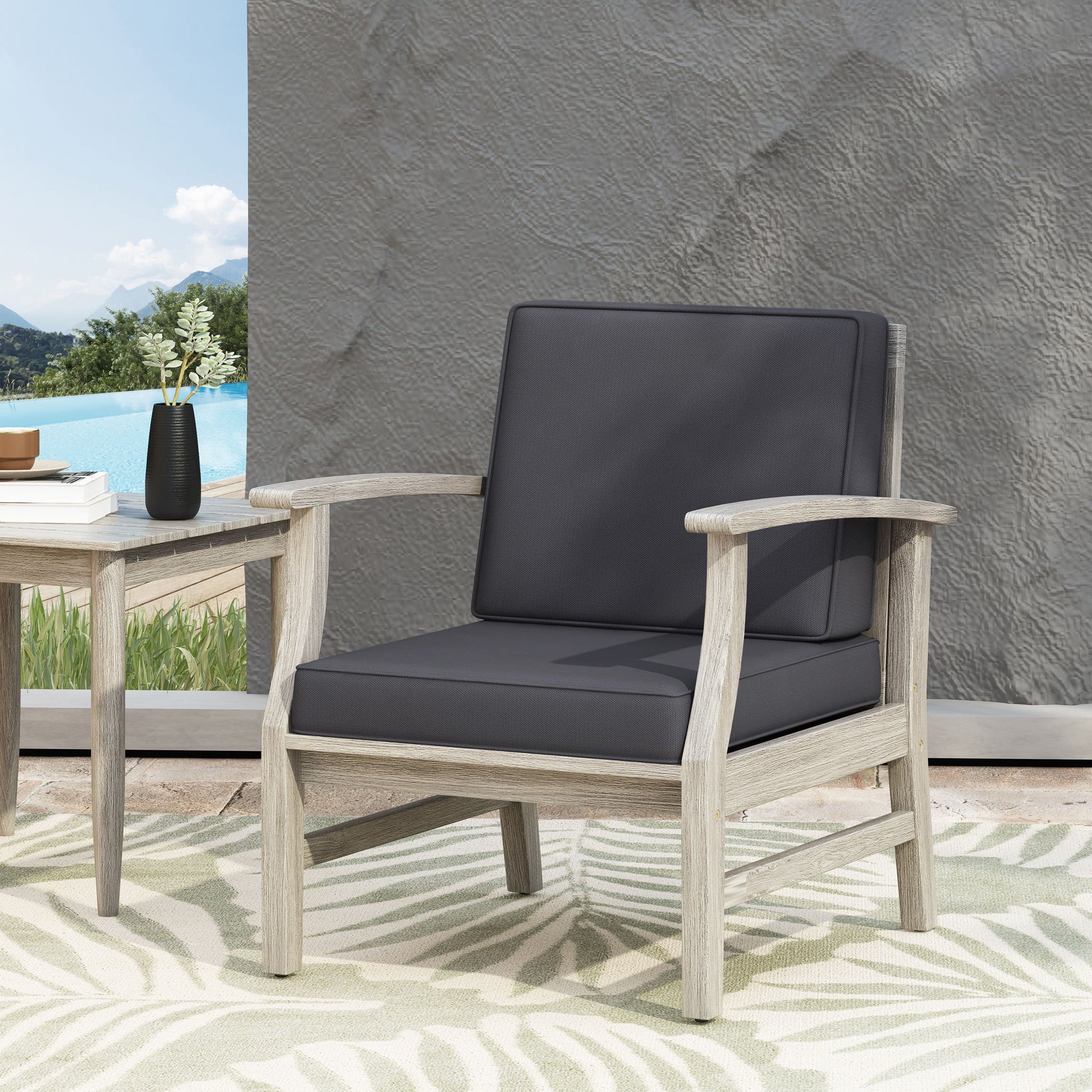 The Perla Contemporary Club Chair with water-resistant gray cushions and a light wood frame sits on a leaf-patterned rug beside a small table with a vase, against a textured gray wall near a pool and mountain backdrop.