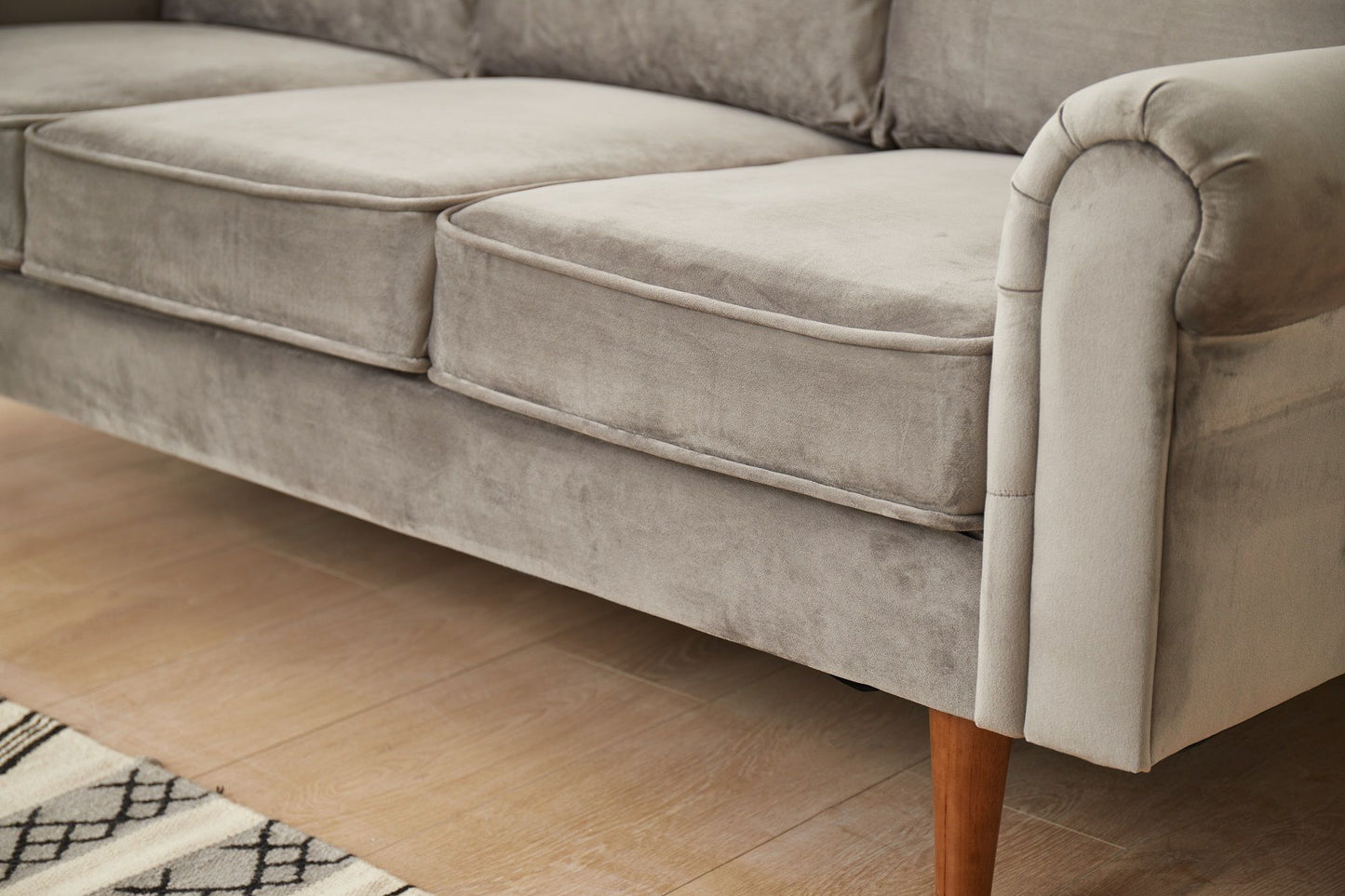 The Graceful Design Plush Comfort Sofa in light gray velvet features three seat cushions and wooden legs, shown on a light wood floor beside a patterned rug.