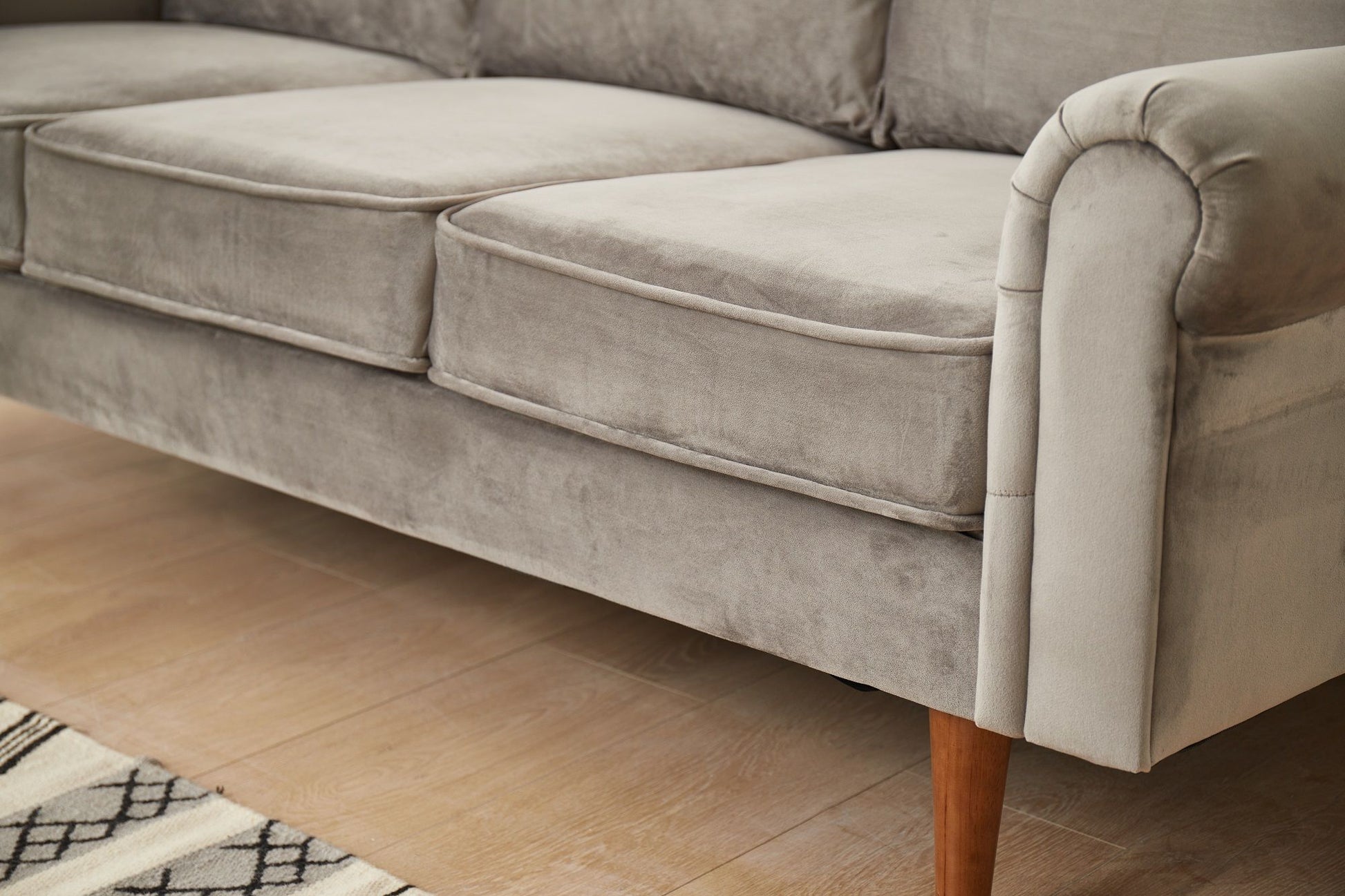 The Graceful Design Plush Comfort Sofa in light gray velvet features three seat cushions and wooden legs, shown on a light wood floor beside a patterned rug.