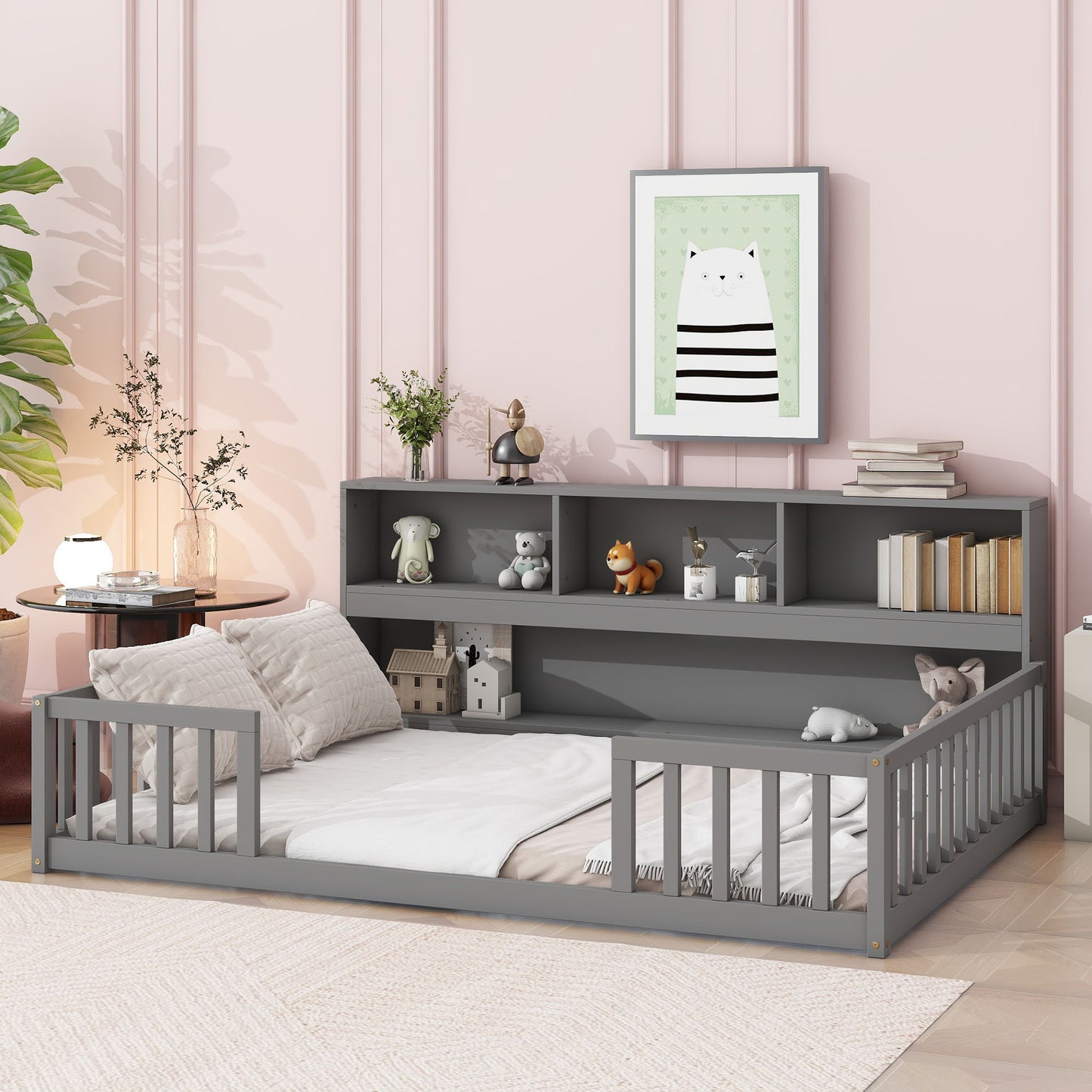 A cozy gray Floor Bed With Side Bookcase, guardrails, and bedding sits against a pink paneled wall. The shelves above display books, toys, and decor. A framed cat print hangs above, with a leafy plant nearby.