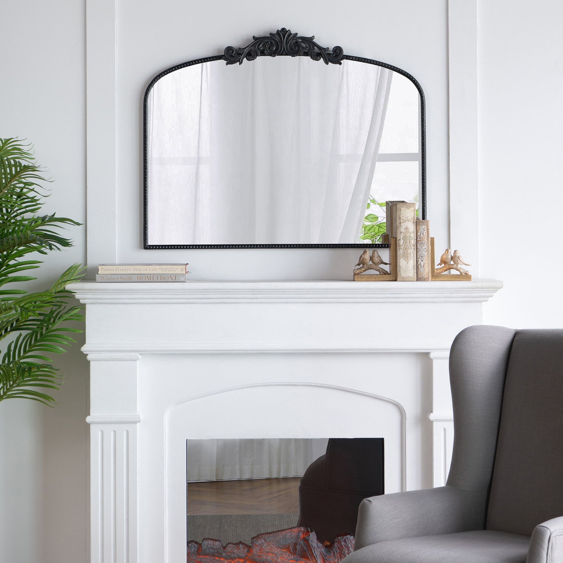 A white fireplace features the Arch Mirror, Baroque Inspired Wall Decor for Bathroom, Bedroom, Living Room above it, flanked by book stacks with decorative bookends. A gray armchair and a green plant sit nearby in the bright space.