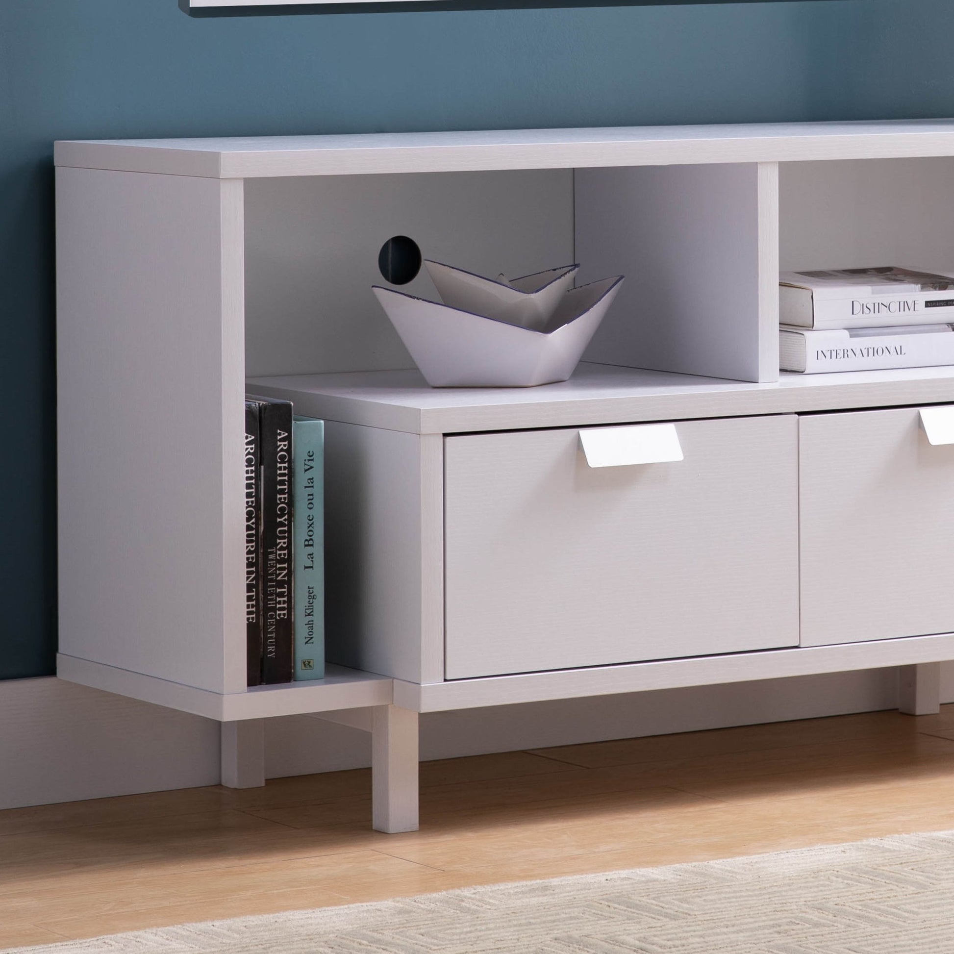 The Modern TV Stand features two shelves and two drawers in white, displaying books, magazines, and a geometric bowl against a blue wall on a light wooden floor.