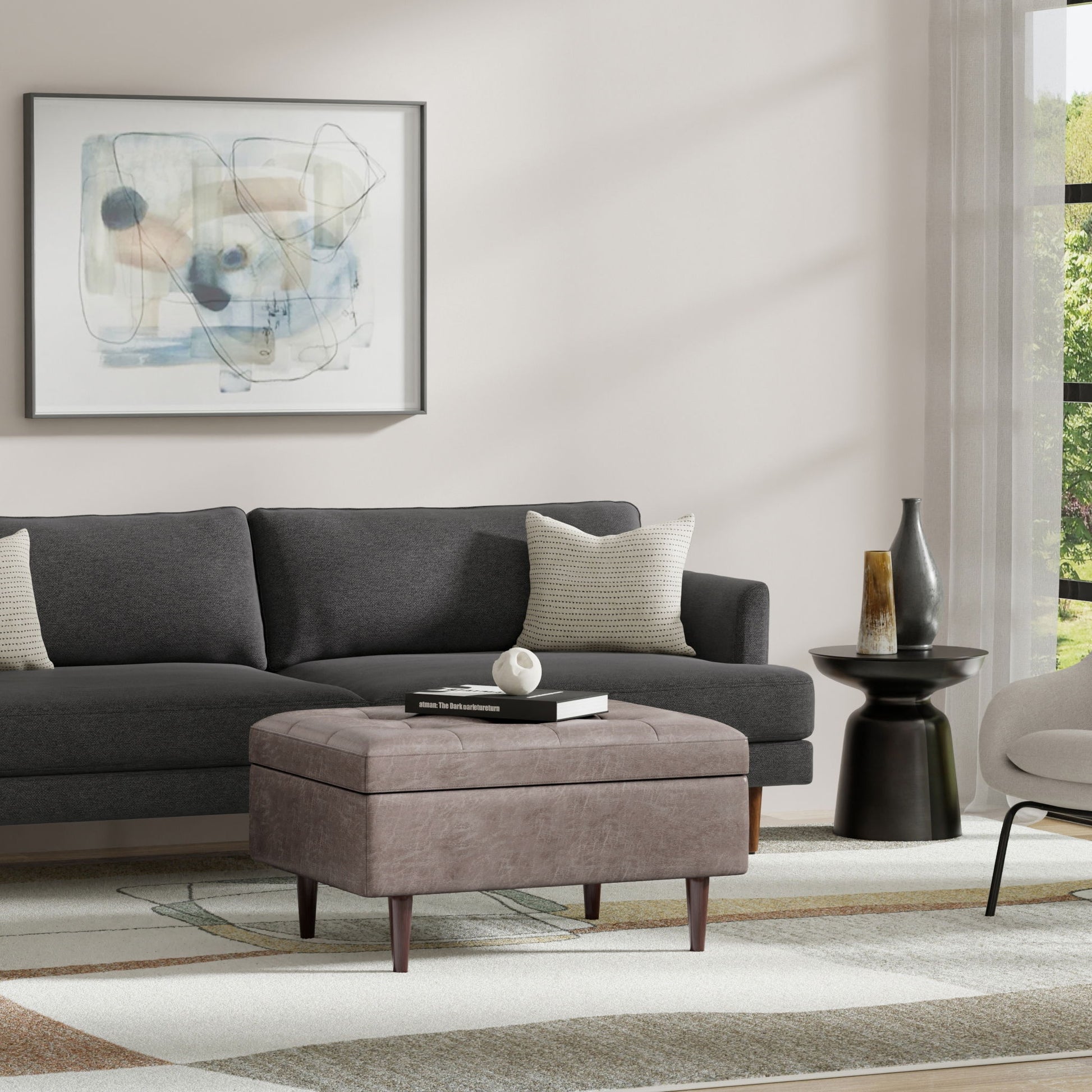 A modern living room with a dark gray sofa, two neutral pillows, an abstract painting, the Shay Large Square Coffee Table Storage Ottoman in mid-century style, a black side table with vases, and sheer curtains letting in natural light.