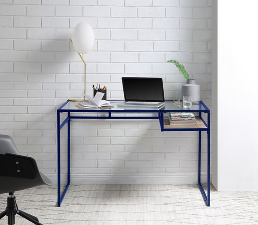 The Yasin - Glass Top Writing Desk with a blue metal frame stands against a white brick wall, holding a laptop, lamp, plant, books, notepad, and pencils. A black chair is partially visible in the foreground.