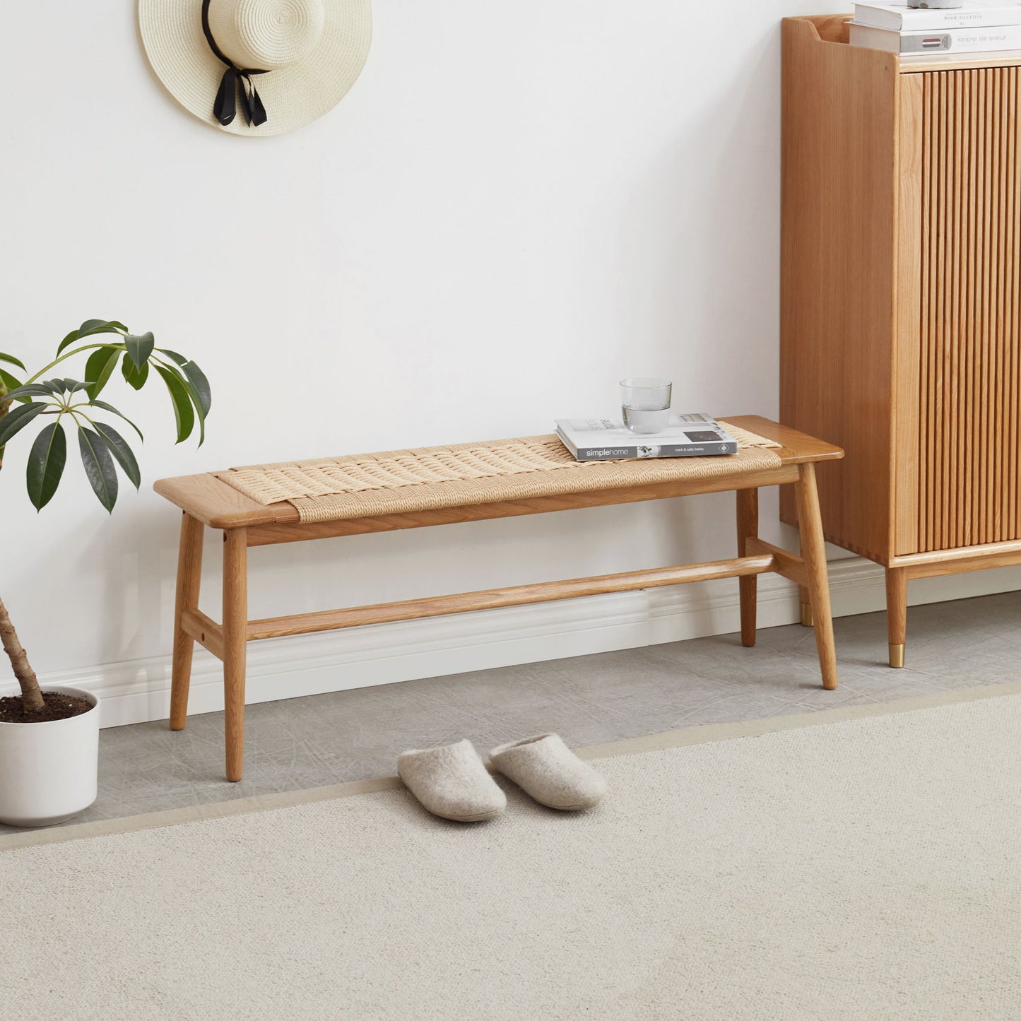A minimalist room features the Woven Design Dining Bench for Dining Room, Bedroom - Natural Oak, topped with a glass of water, books, and a tray. Nearby are gray slippers, a potted plant, a cabinet, and a straw hat on a white wall.