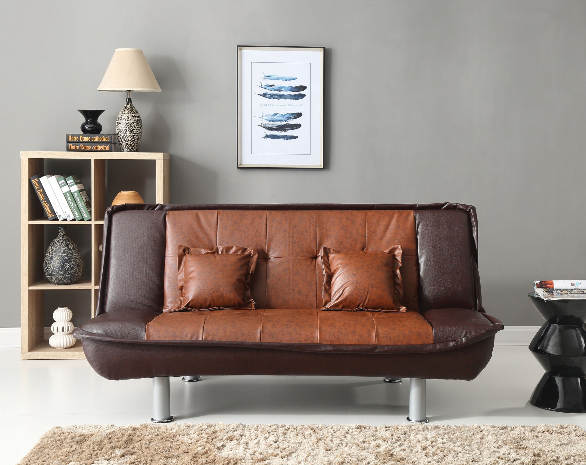 A modern living room showcases a two-tone Lionel sofa bed, featuring brown upholstery with two matching cushions. A wooden shelf holds books and decor, paired with a black and cream lamp, while framed artwork adorns the gray walls above a light-colored floor.