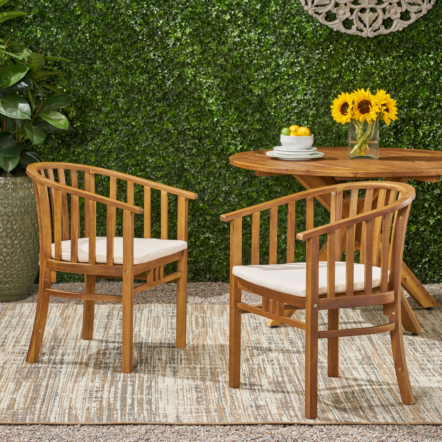 Two Outdoor Wooden Dining Chairs With Cushions (Set of 2) sit on a rug outdoors beside a wooden table with sunflowers and lemons. A green hedge and potted plant are in the background.