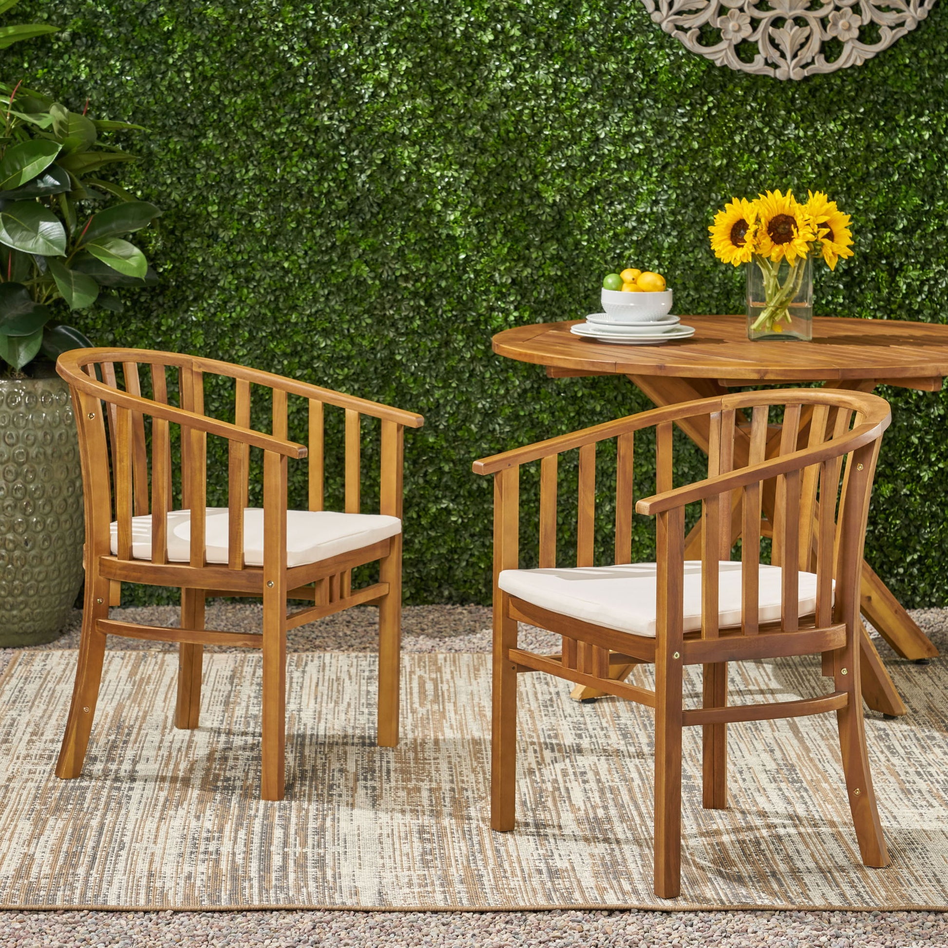 Two Outdoor Wooden Dining Chairs With Cushions (Set of 2) sit on a rug outdoors beside a wooden table with sunflowers and lemons. A green hedge and potted plant are in the background.