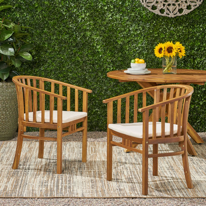 Two Outdoor Wooden Dining Chairs With Cushions (Set of 2) sit on a rug outdoors beside a wooden table with sunflowers and lemons. A green hedge and potted plant are in the background.