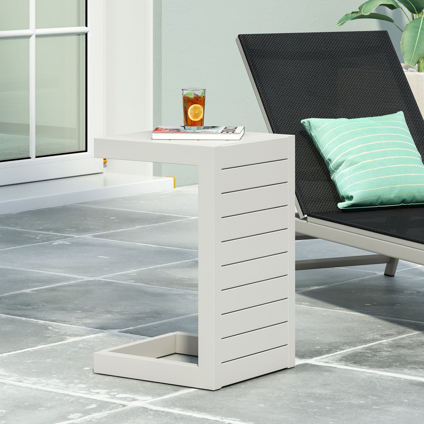 The Aluminum Outdoor C-Shaped Side Table for Patio, featuring a modern white design with horizontal grooves, stands on a gray tiled patio beside a black lounge chair and holds a drink with orange slices and ice, next to a book.